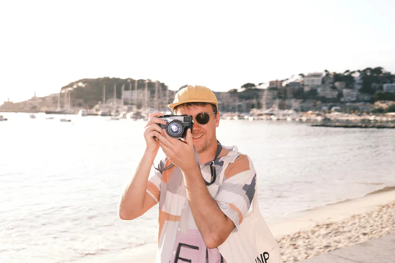 Man in a yellow cap and sunglasses holding a camera by a seaside beach at sunset.