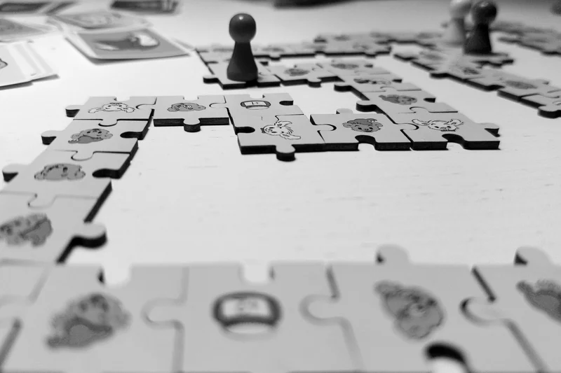 Black-and-white jigsaw puzzle pieces laid out on a table with two pawn-like game pieces in between.