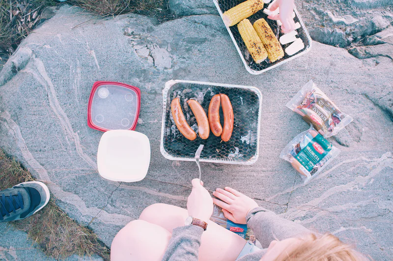 Picnic setup on rocks with hot dogs in a grill basket, corn on the cob, and hands preparing food.