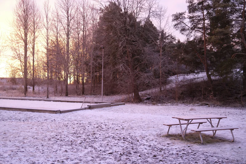 Snowy clearing with a wooden bench and covered picnic area near a sandbox, surrounded by winter trees.