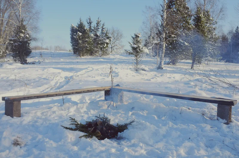 Wooden log bench around a campfire with a pile of burning wood in a snowy winter field.