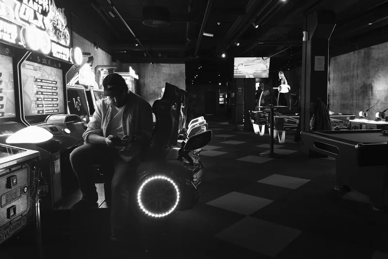 Man in a dark arcade room sits at a pinball machine while other gaming machines line the aisle.