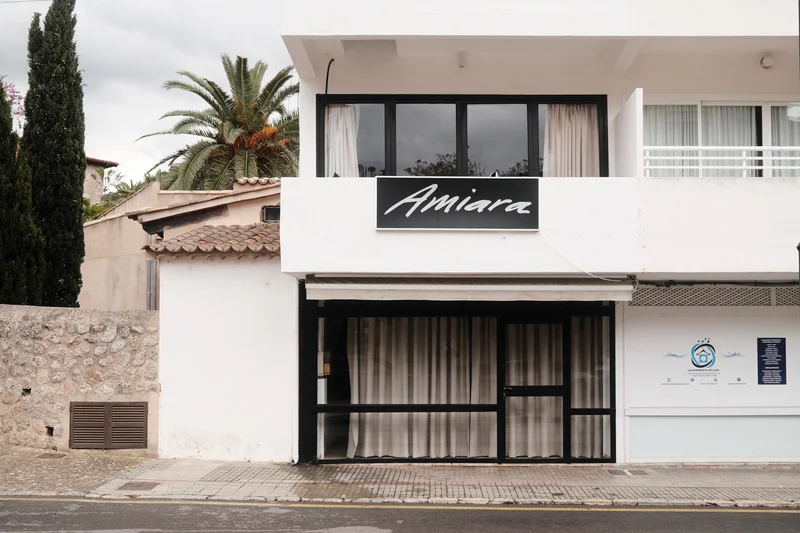 White building with a black sign reading “Amiara” above an entrance and windows, with palm trees nearby.