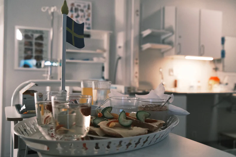 Tray on a hospital table with Swedish flag, drinks, and open sandwiches in glass bowls.