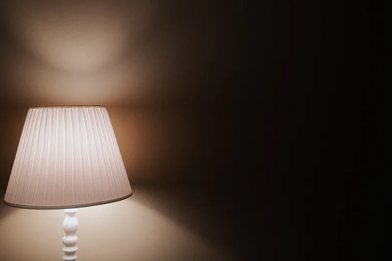 Table lamp with a white pleated shade on the left side of a dim, blurred room.