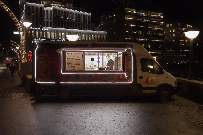 Night street scene with a food truck displaying a lit menu board in front of office buildings.