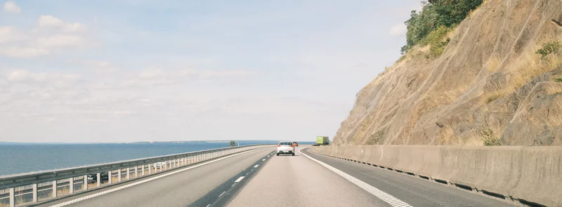 Two-lane highway over the sea with guardrails, a few vehicles, and a steep rocky hillside on the right.