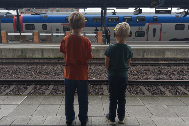Two young boys in casual clothes stand on a train platform facing a blue and white passenger train.