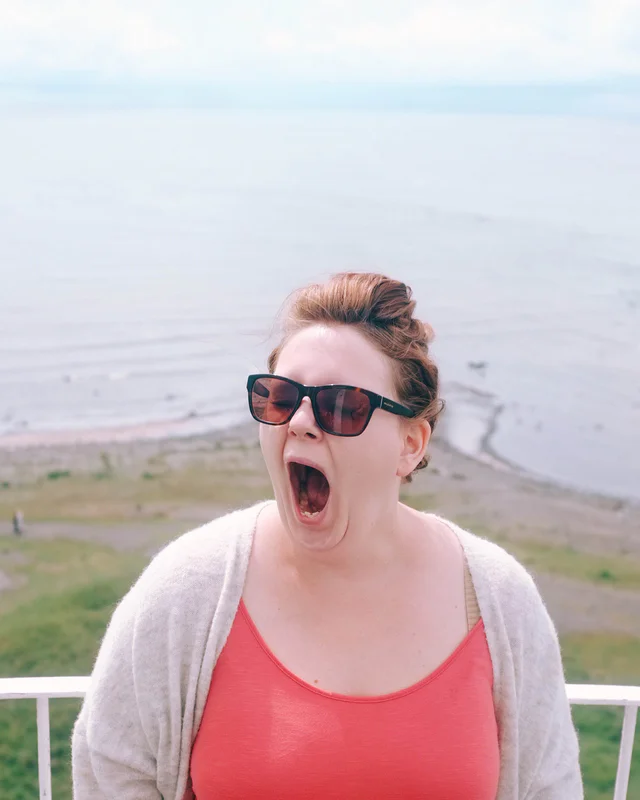 Woman wearing sunglasses yawns with mouth wide open outdoors by a mountainous landscape.