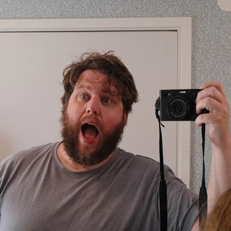 Bearded man taking a selfie with a black camera, mouth open, in a mirror.