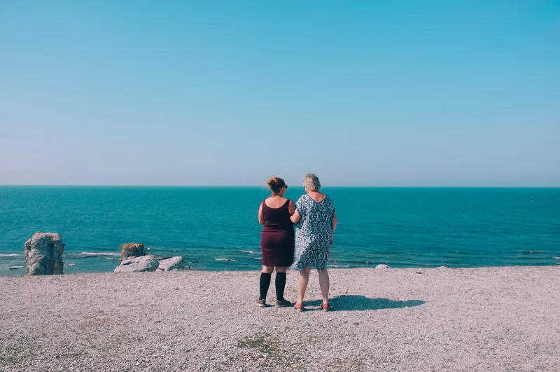 Two adults stand on a rocky beach facing the ocean under a clear blue sky.