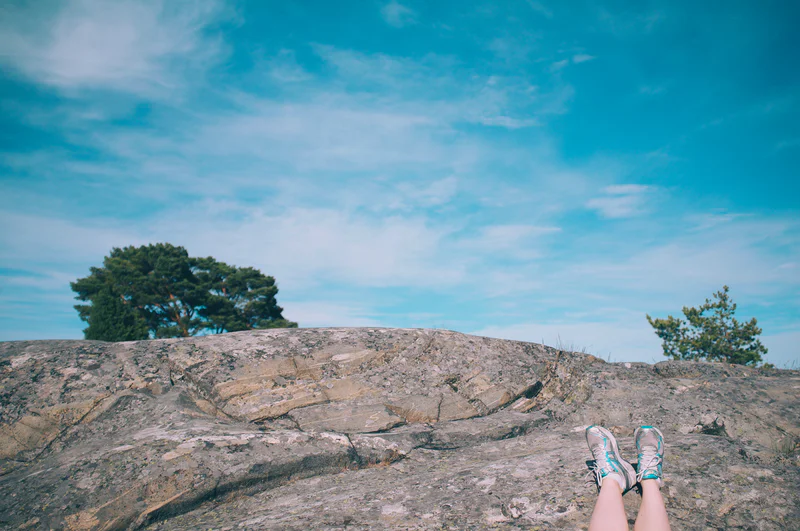 Person’s legs in hiking shoes at rocky overlook with trees and blue sky in the distance.