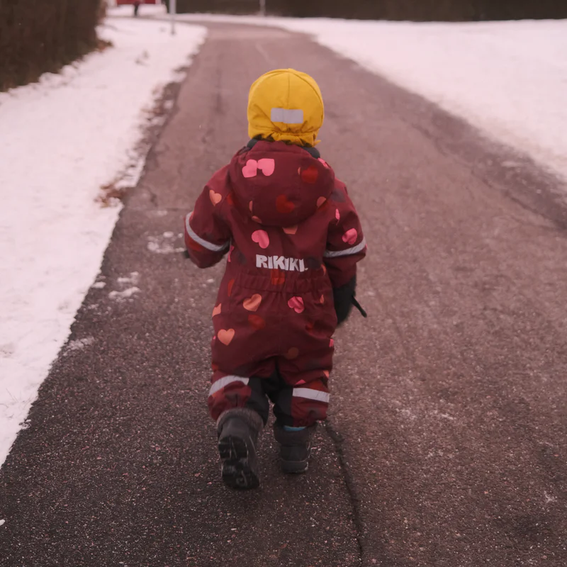 Small child in a maroon snowsuit with a yellow hood walks on a snowy street, viewed from behind.