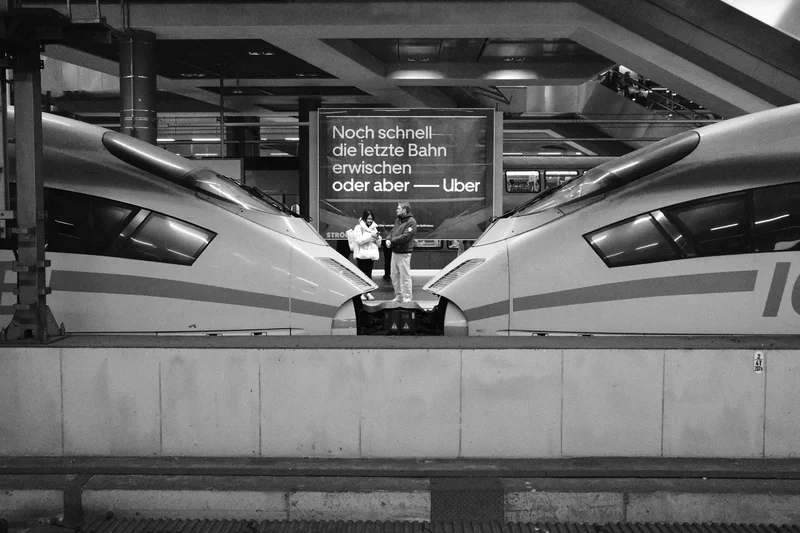 Two people stand between two parked high-speed trains in a station, with German text on a sign above.