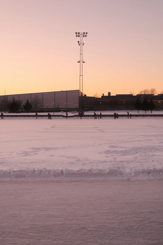 Snow-covered outdoor field at dusk with a tall floodlight pole and people scattered across the ice.