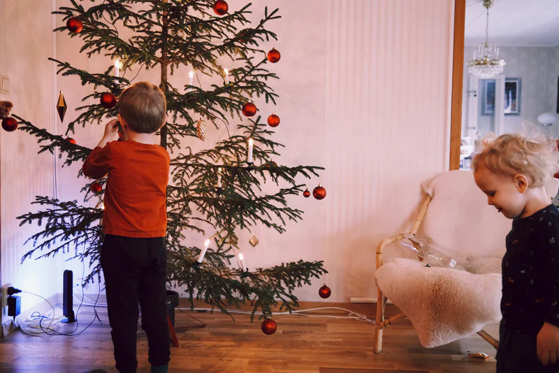 Two children stand near a decorated Christmas tree with red ornaments in a living room.