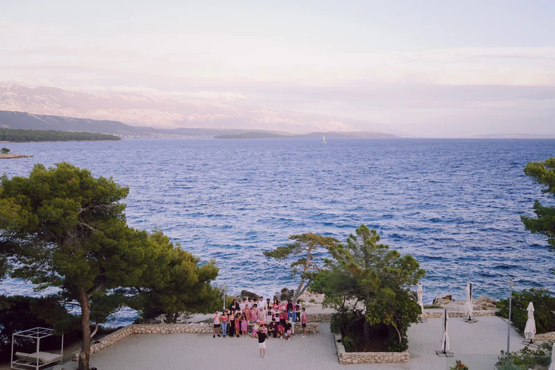 Group photo on a seaside terrace overlooking calm blue water and distant hills at dusk.