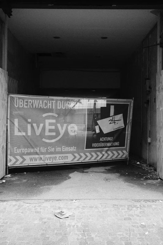 Abandoned corridor with a broken poster reading “Liveye,” “Überwachung,” and “Achtung,” plus a “24” sign.