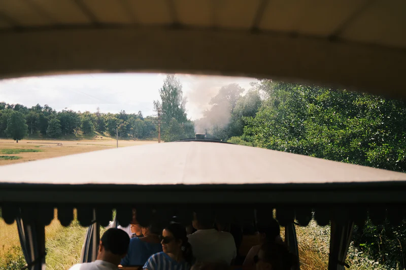 View from inside a train carriage toward an outdoor track, passengers seated, with trees and steam visible ahead.