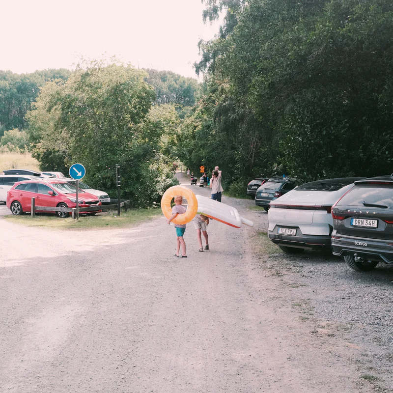 People stand near a parked cars lot on a gravel path, with an orange inflatable ring on a child.