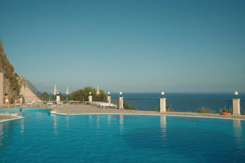 Outdoor hotel pool with sun loungers, umbrellas, and a seaside view under a clear sky.