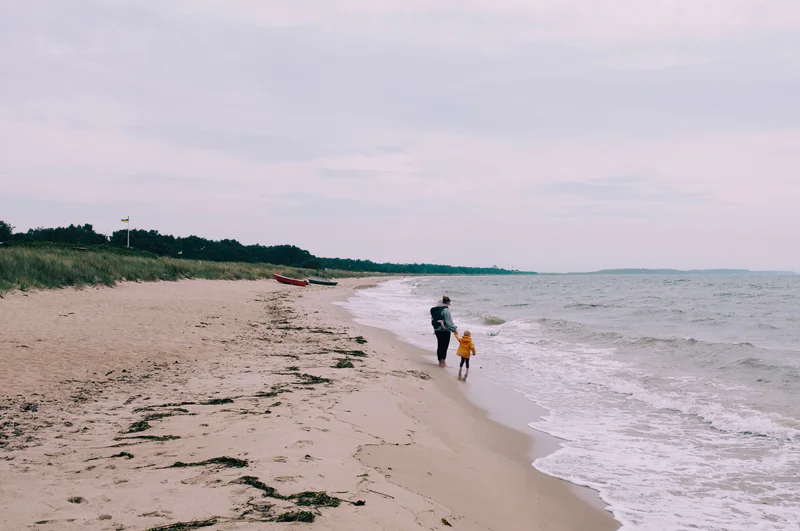 Two people walk along a sandy beach holding hands as waves roll in at dusk, with a small boat in the distance.