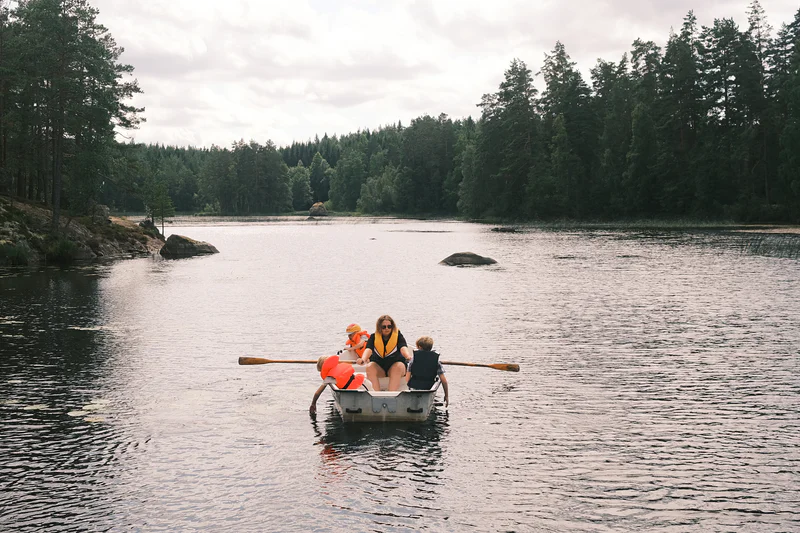 A woman and three children ride in a small rowboat on a calm lake near forested shores.