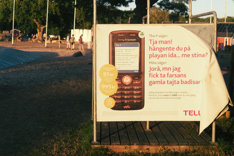 Billboard advertising a mobile phone in a grassy campsite at dusk, with a blue tarp and people in the background.