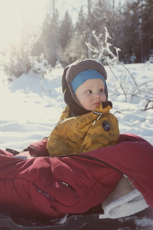 Baby wearing a blue hat and yellow winter jacket sits in a red sled outdoors in snowy woods.
