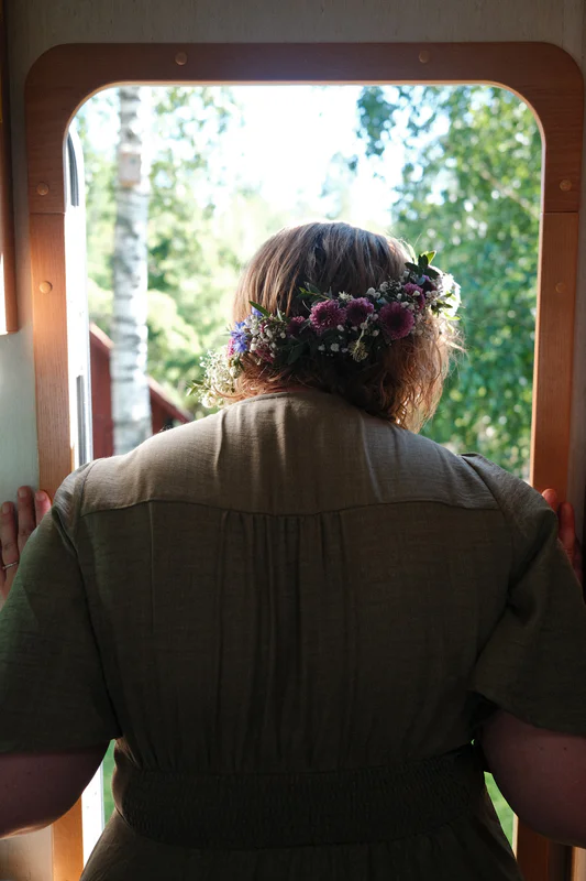 Person seen from behind with a purple flower crown, standing by an open doorway with trees outside.
