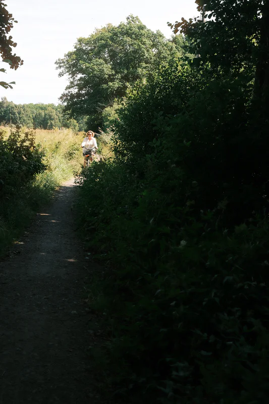 Woman on a bicycle riding along a narrow dirt path through tall green trees.