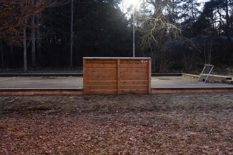 Brown wooden fence gate with a light pole behind it in a leaf-covered outdoor yard.