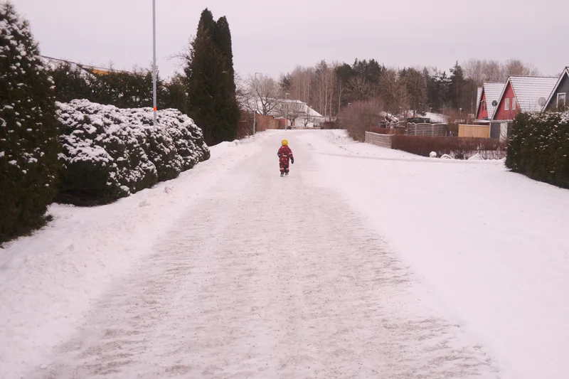 Child in colorful snowsuit runs down a snowy residential driveway toward houses and trees.