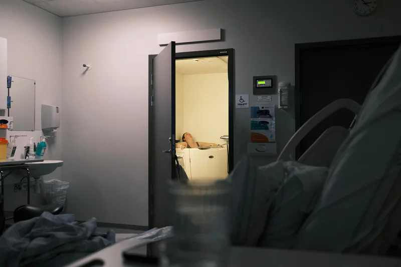 Woman sits in a bathtub in a hospital room, seen through an open doorway.
