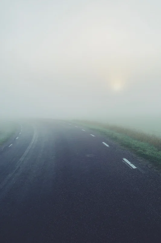 Road stretching into fog with dashed lane markings and a distant glare in the sky.