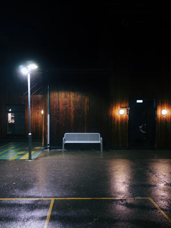 Nighttime parking lot with streetlights, wet asphalt, and a bench against a dark wooden wall.