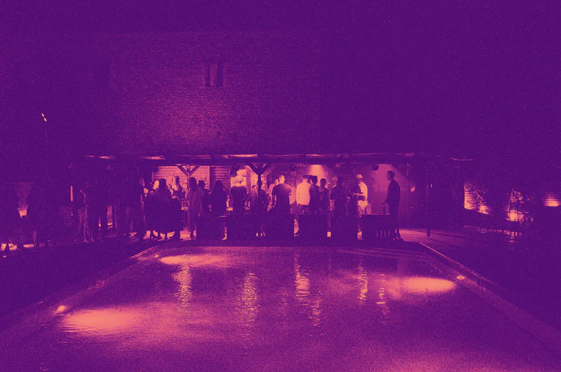 People stand and walk outside a pavilion beside a lit outdoor swimming pool at night.