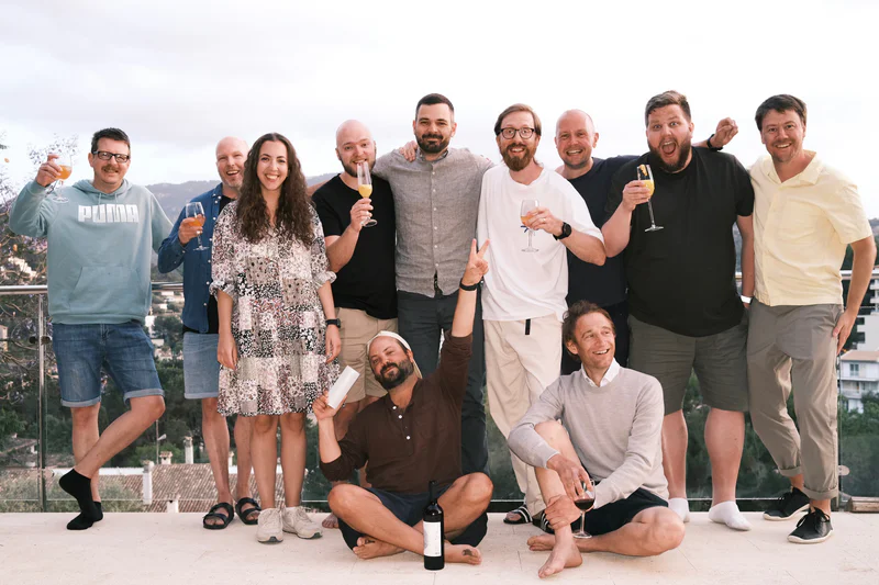 Group of eight adults posing outdoors on a terrace, holding champagne flutes and a bottle.