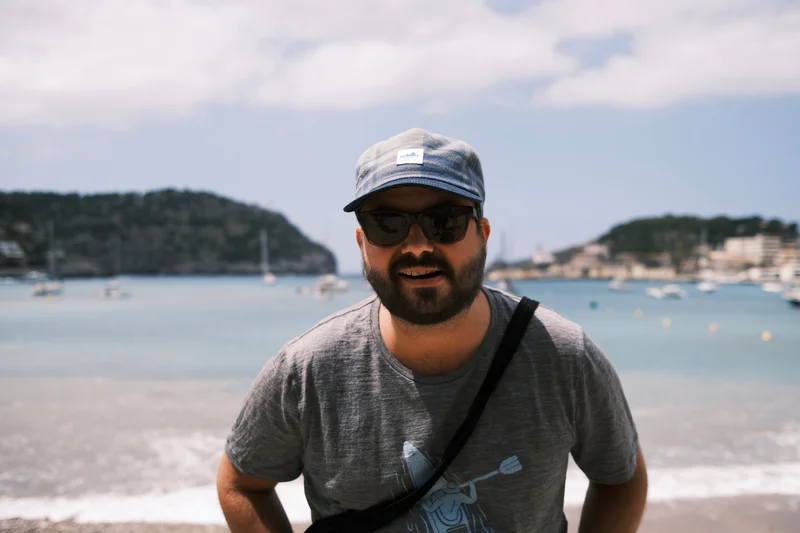 Man wearing a cap and sunglasses, standing on a beach with water and hills in the background.