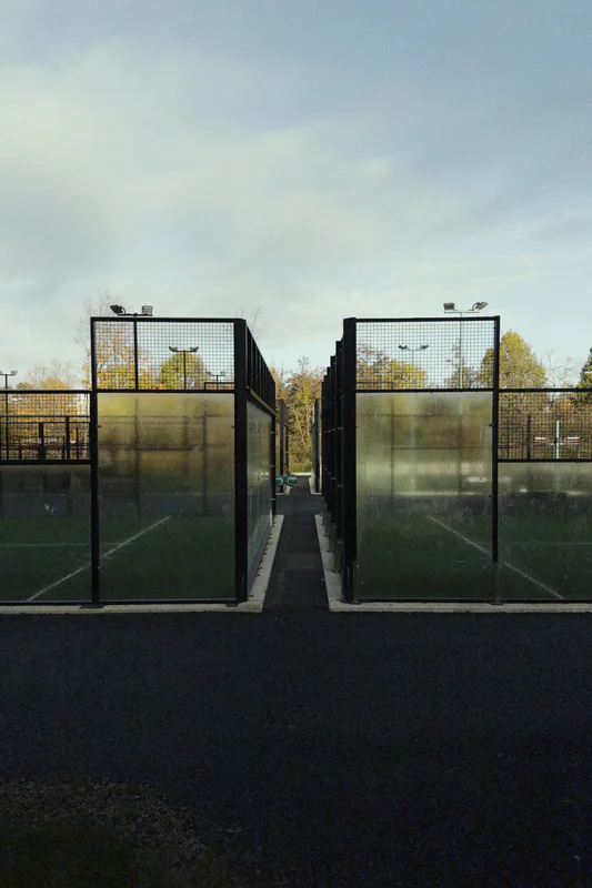 Outdoor tennis court with two closed metal gates and a fence-lined walkway under a cloudy sky.