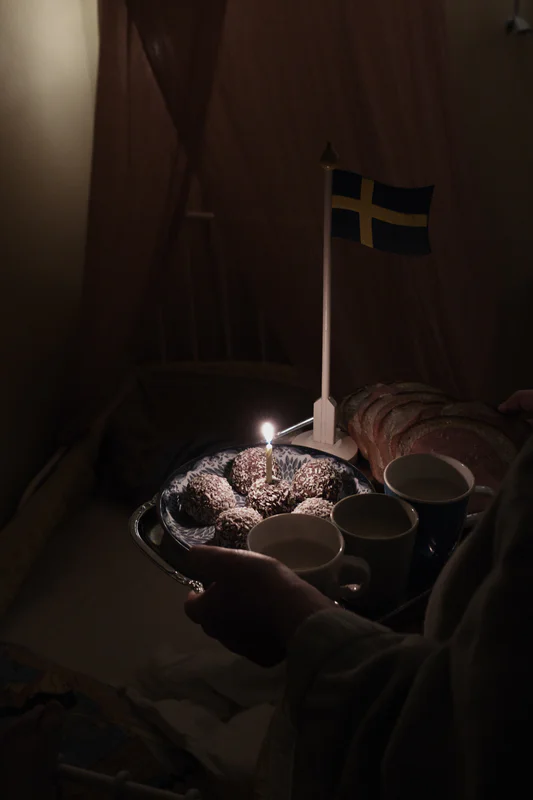 Person holds a tray of candles, cups, and decorated buns with a small Swedish flag on a lit cake in dim light.