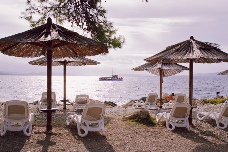 Two thatched beach umbrellas and empty lounge chairs on a rocky shore beside the ocean.