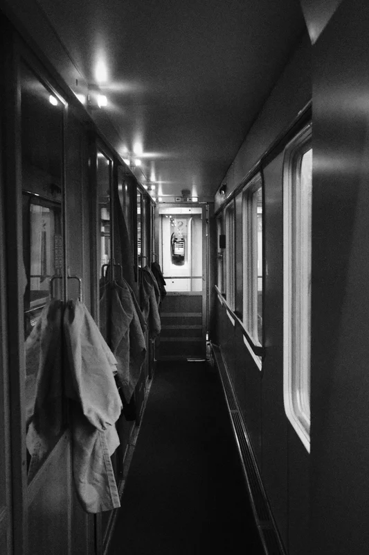 Narrow monochrome train corridor with rows of seats and hanging jackets under bright ceiling lights.