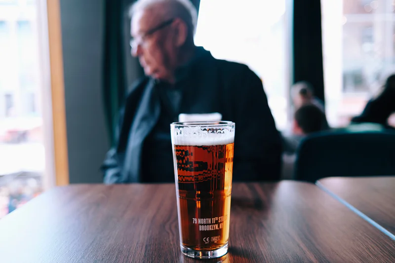 Tall pint glass of amber beer with foam on a wooden table, with a blurred man seated behind it.