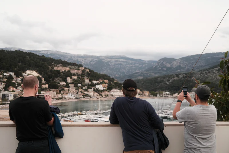 Three people stand on a terrace photographing a harbor town and mountains under an overcast sky.