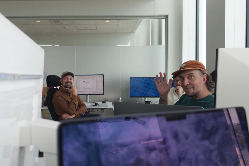 Three men in a bright office, sitting at desks and talking behind computer monitors.