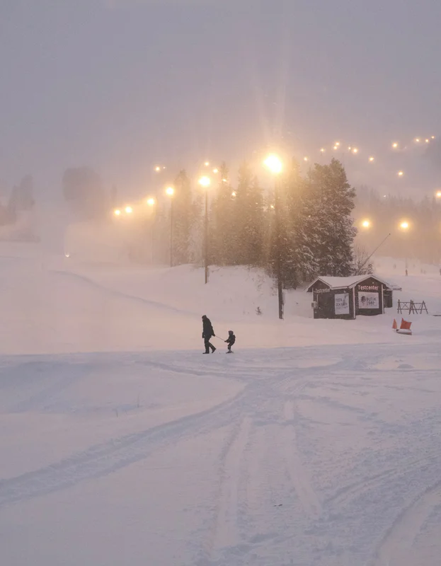 Snowy ski slope at dusk with glowing streetlights, trees, and a small building near a trail.