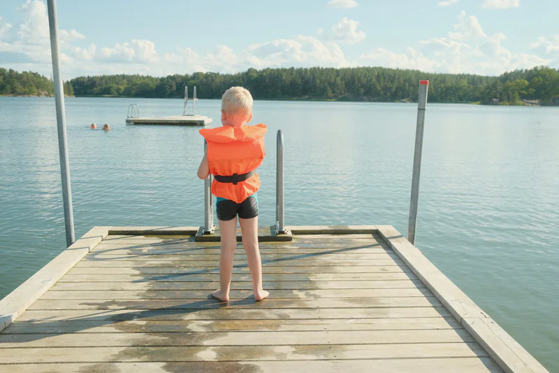 A child with blond hair stands barefoot on a wooden dock, wearing an orange life jacket, overlooking a lake.