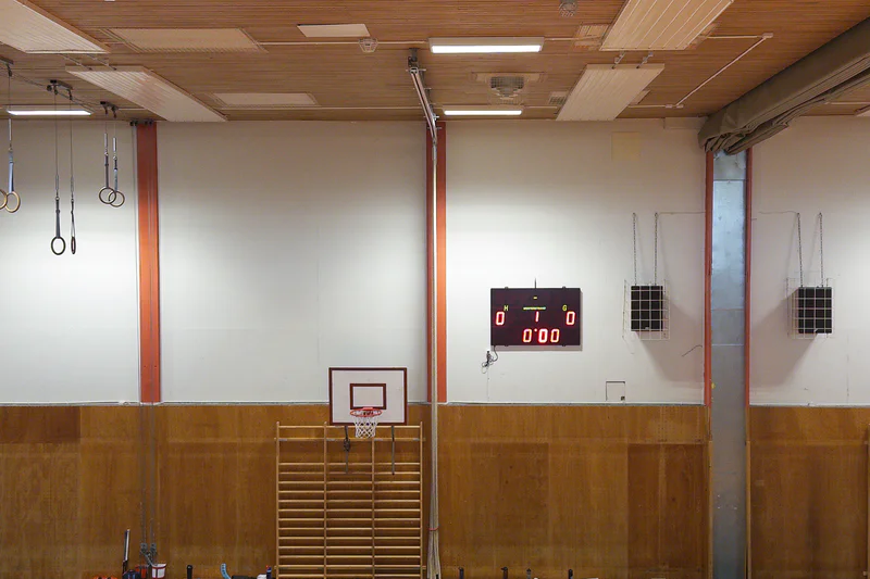 Indoor basketball court with a hoop, scoreboard on the wall, and wall-mounted rings and lights.