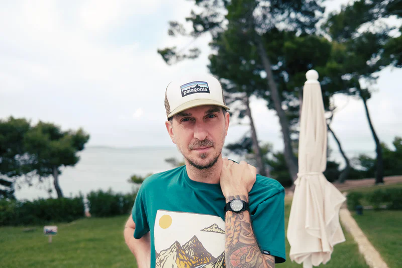 Man in a green shirt and Patagonia cap standing outdoors near a seaside park with a white umbrella nearby.